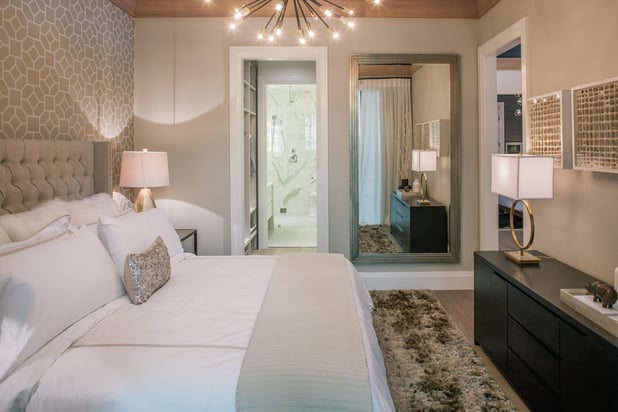 This bedroom has white and tan bedding with a furry, brown rug at the end of the bed and a lighting fixture hanging in the center of the room.