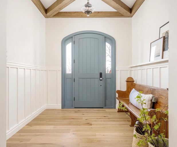 An entryway with a blue door, white walls and moulding, a repurposed brown church pew used as a bench and some picture frames resting on moulding above the pew. 