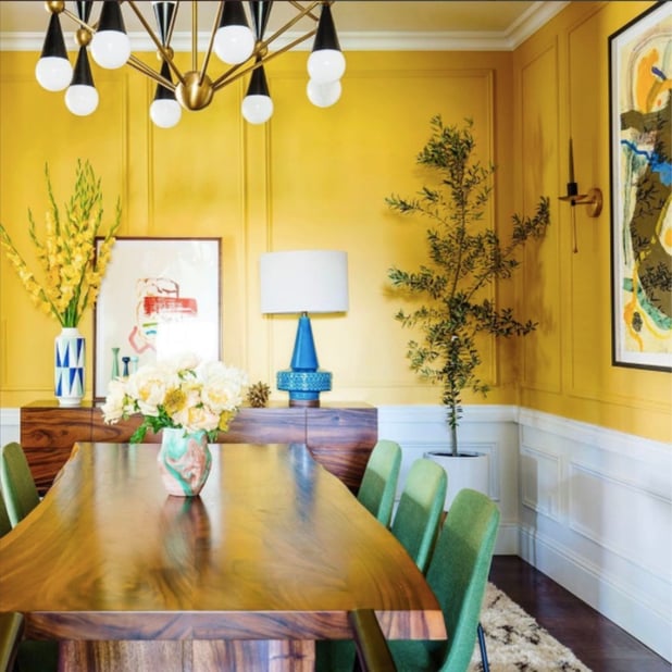 Modern dining room with yellow walls, white trim, green dining chairs, a blue lamp, yellow flowers, a green tree and colorful artwork.