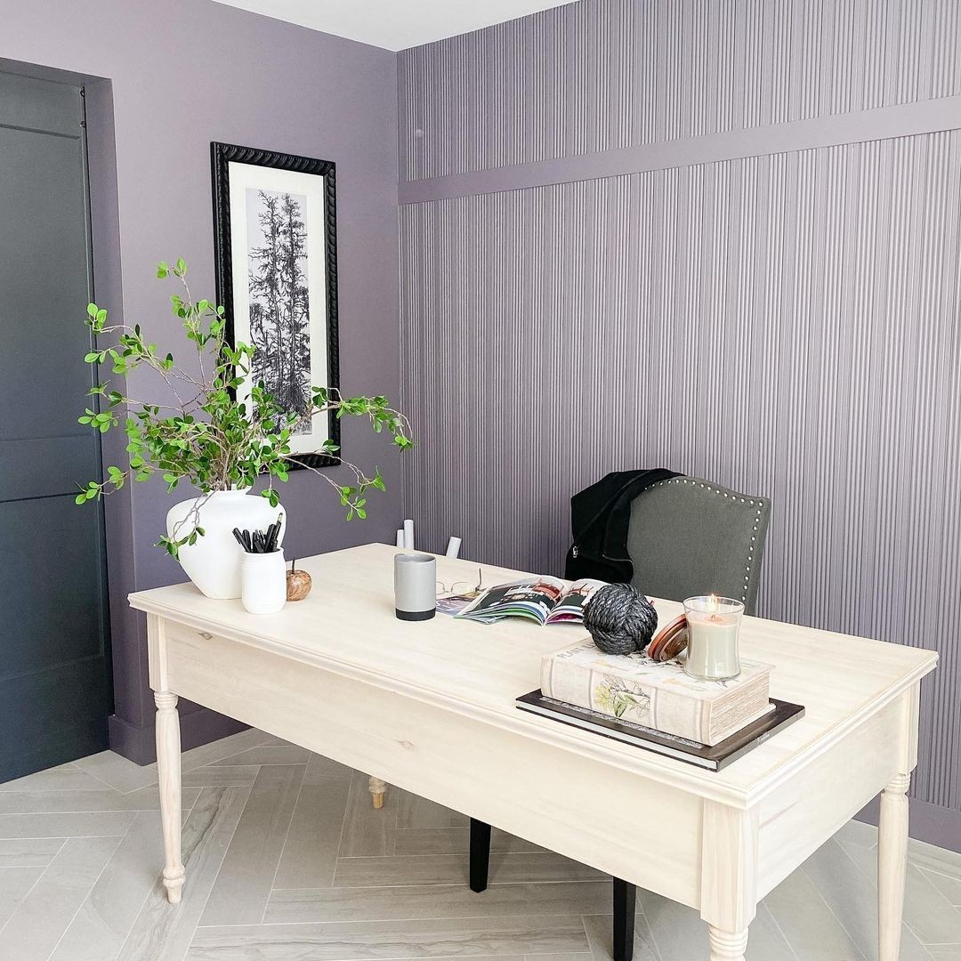 This office space has a white wooden desk, a grey upholstered chair, purple moulding and walls, and grey, patterned panel flooring.